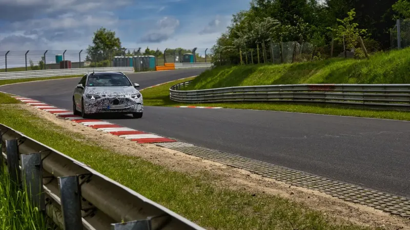 A camouflaged sports car speeds around the Nürburgring race track on a sunny day.