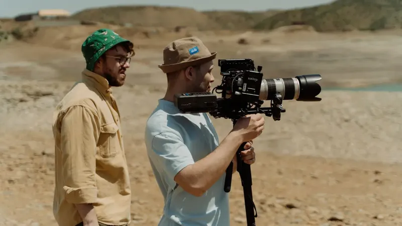 Two filmmakers operating a professional video camera outdoors in a rocky landscape.
