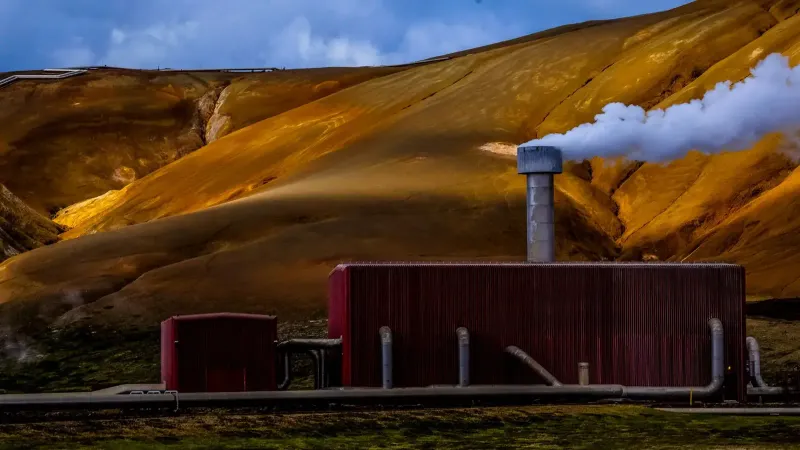 Planta de energía geotérmica roja que emite vapor contra un dramático telón de fondo montañoso de color amarillo-marrón.