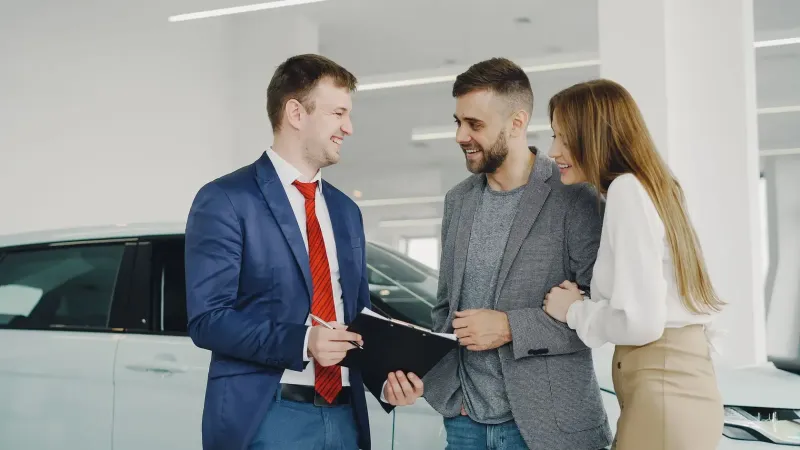 Una pareja sonriente compra un automóvil nuevo a un vendedor seguro dentro de un concesionario de automóviles moderno.