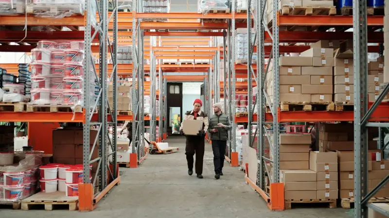 Dos trabajadores manejan un paquete en un amplio almacén rodeado de estantes repletos de cajas y productos.