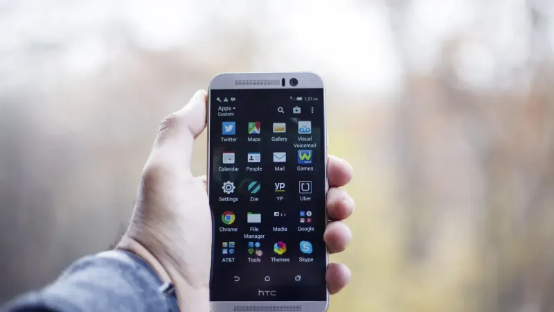 A close-up of a hand holding an HTC smartphone with visible apps on the screen, in an outdoor setting.
