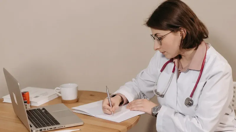 Doctora con estetoscopio escribiendo en el escritorio con laptop y notas.