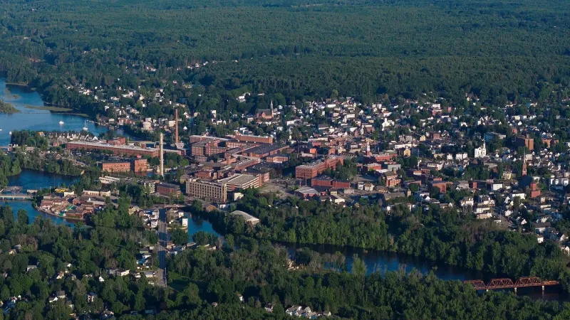 Vista aérea del centro de Biddeford, Maine, con antiguos edificios de ladrillo y chimeneas junto a un río, casas entre árboles y un puente metálico rojizo.