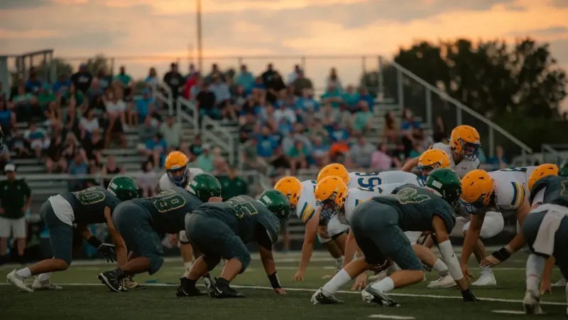 Teams face off in an intense high school football game as the sun sets, adding drama to the scene.