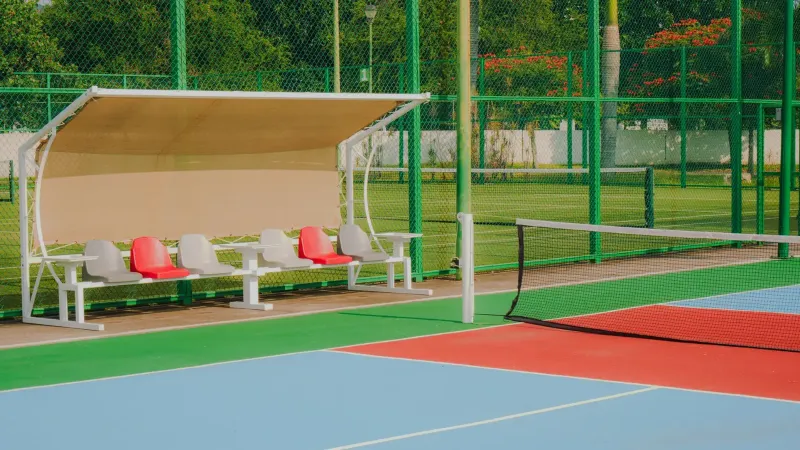 Vibrant tennis court featuring colorful flooring, seating benches, and lush greenery, captured outdoors in daylight.