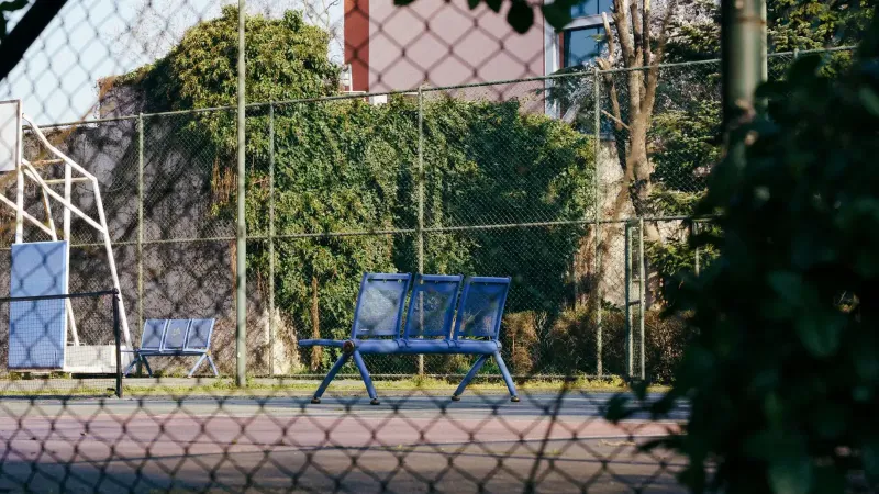 A serene view of an empty sports court with blue benches, surrounded by greenery and fencing.