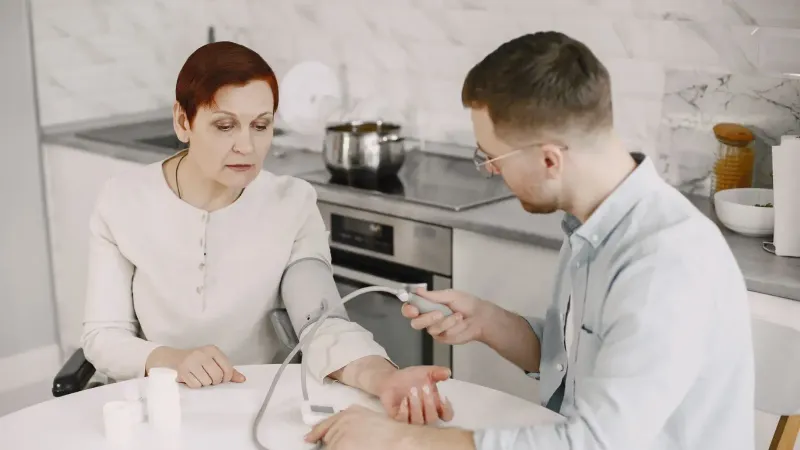 Adult man monitoring elderly woman's blood pressure at kitchen table using sphygmomanometer.