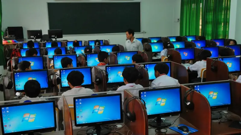Students learning in a computer lab with multiple monitors and a teacher instructing.