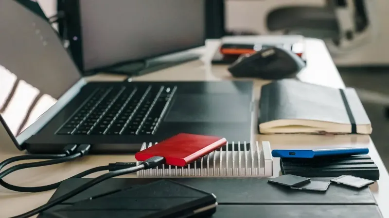 Escritorio de oficina moderno con computadora portátil, unidades externas y varios accesorios tecnológicos para un flujo de trabajo eficiente.