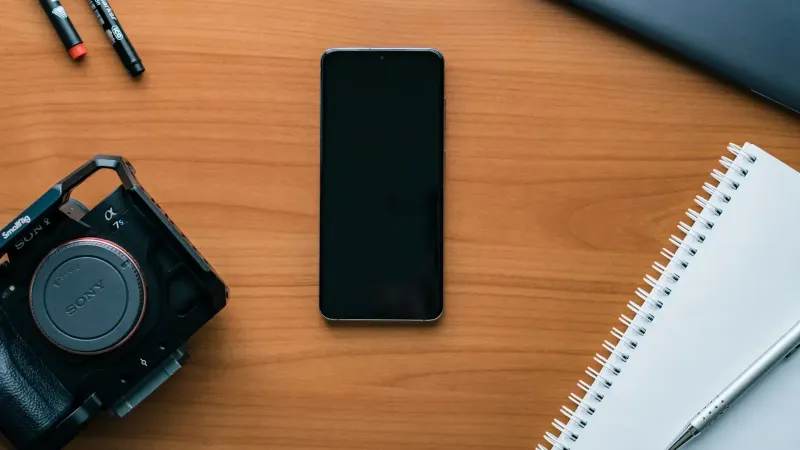 Una ordenada y plana de un espacio de trabajo moderno con un teléfono inteligente, cámara, bolígrafos y portátil.