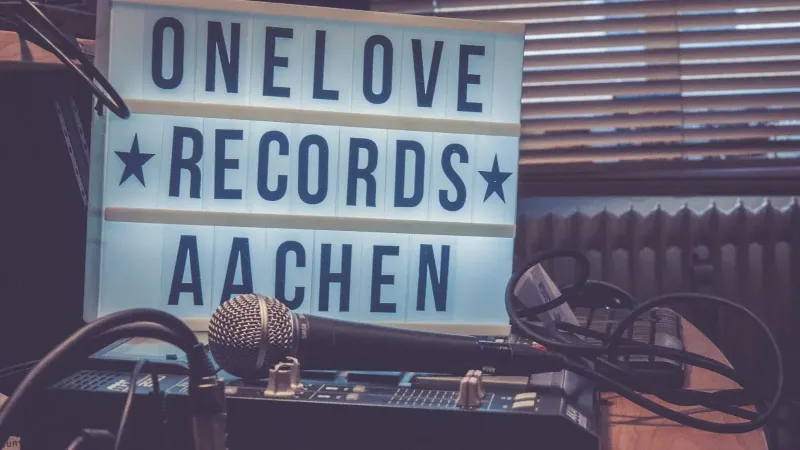 A warm-toned studio featuring a microphone, audio mixer, and lightbox signage reading 'One Love Records Aachen'.
