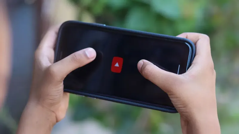 Close-up de manos sosteniendo un smartphone mostrando un icono de aplicación de vídeo al aire libre.