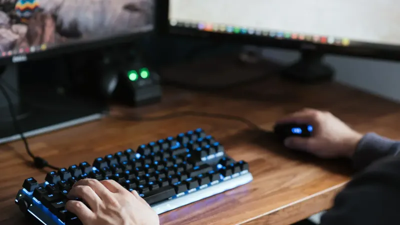 High angle crop male in casual clothes sitting at desk and browsing contemporary computer while typing on backlit keyboard and using mouse