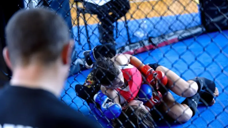 Two women competing in a mixed martial arts training session inside a gym cage with a coach observing.
