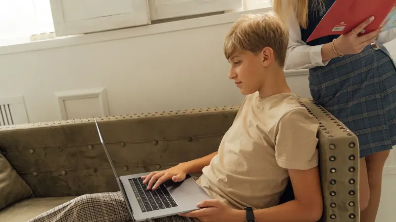 A teenager relaxing on a couch with a laptop, engaging in online learning indoors.