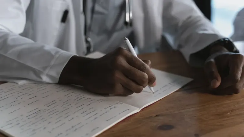 Primer plano de un médico escribiendo notas con un bolígrafo en un cuaderno médico en un escritorio de madera.