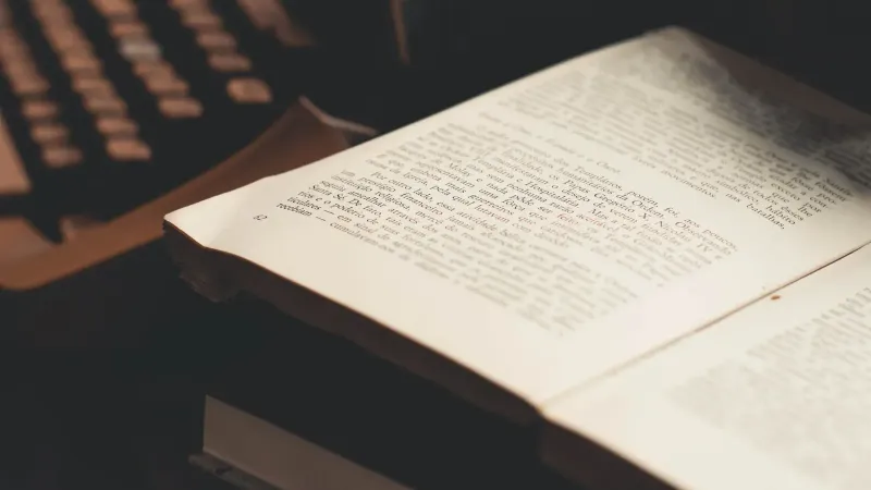 An open book with visible text in front of an antique typewriter, evoking a vintage literary theme.
