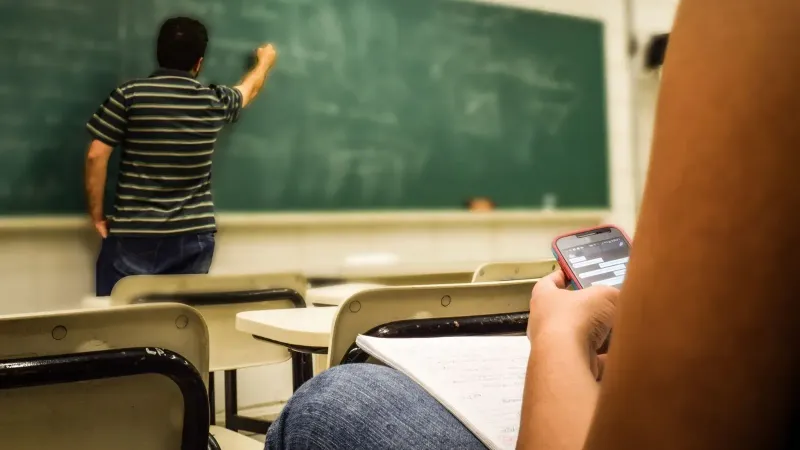 Student texting in a classroom while teacher is writing on the blackboard.