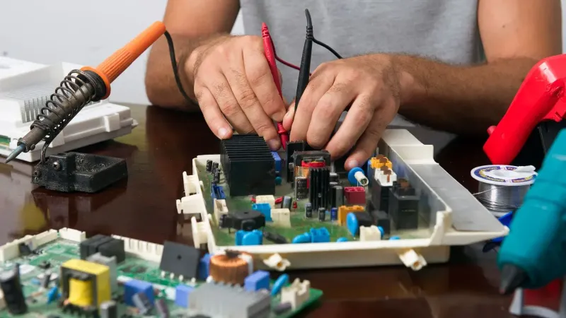 Man working on an electronic circuit board with tools on a table, showing close-up of hands and components.