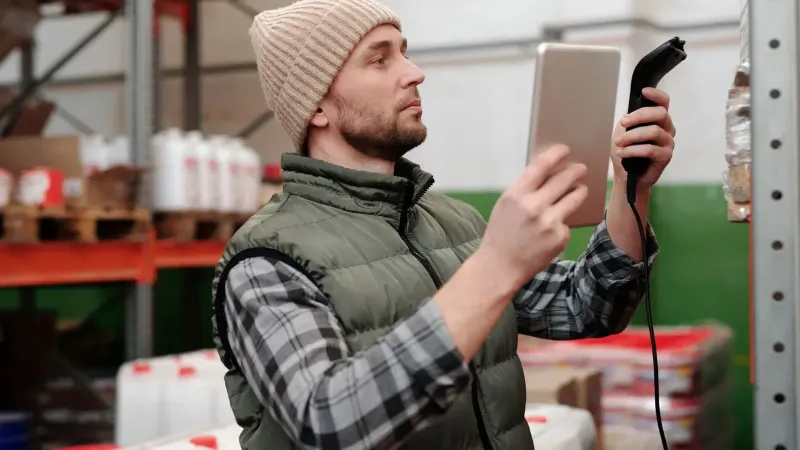 Hombre en gorro y chaleco usando un escáner en un almacén para control de inventario.