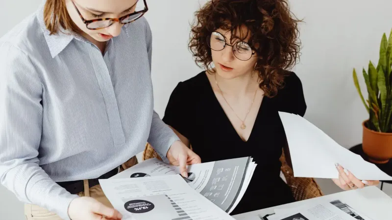 Two women review and discuss financial documents in an office setting, highlighting teamwork and analysis.