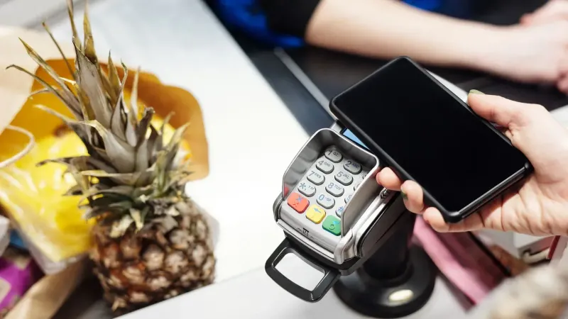 Close-up of a customer using a smartphone for contactless payment at a retail checkout with a pineapple on the counter.
