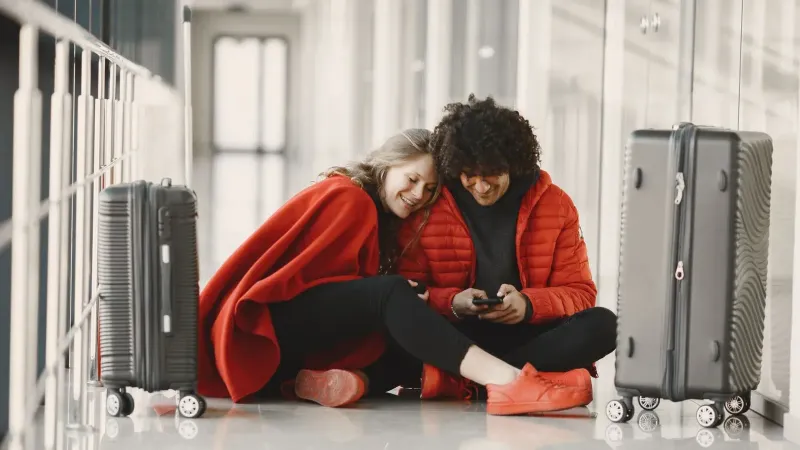Feliz pareja sentada en el piso del aeropuerto con equipaje, compartiendo un momento con smartphone.