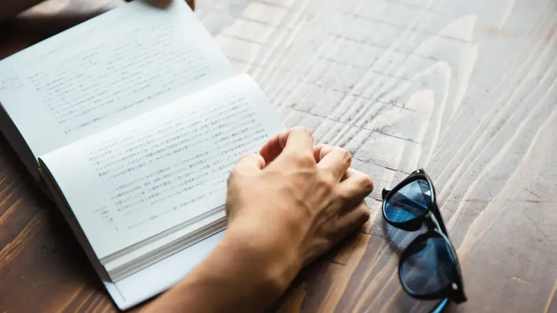 Persona anónima de alto ángulo sentada en la mesa con elegantes gafas de sol azul oscuro y leyendo interesante libro