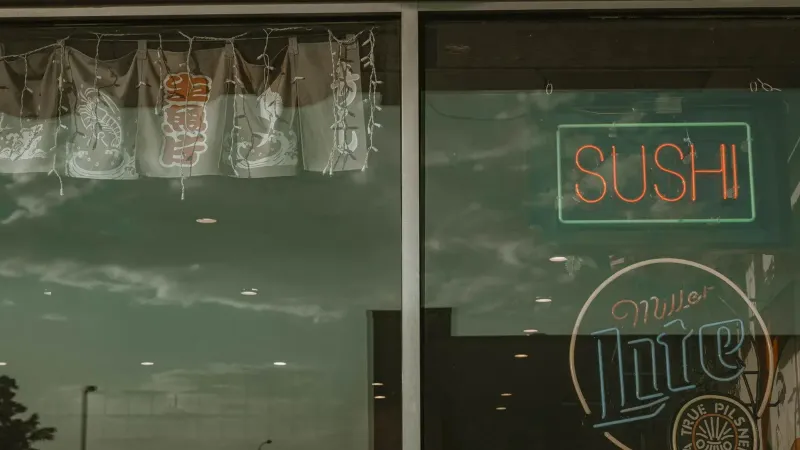 Signo de sushi de neón y reflexión en la ventana del restaurante con cielo nublado.