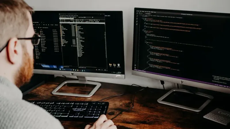 Desarrollador de software que trabaja en código con monitores duales en un entorno de oficina en el hogar.