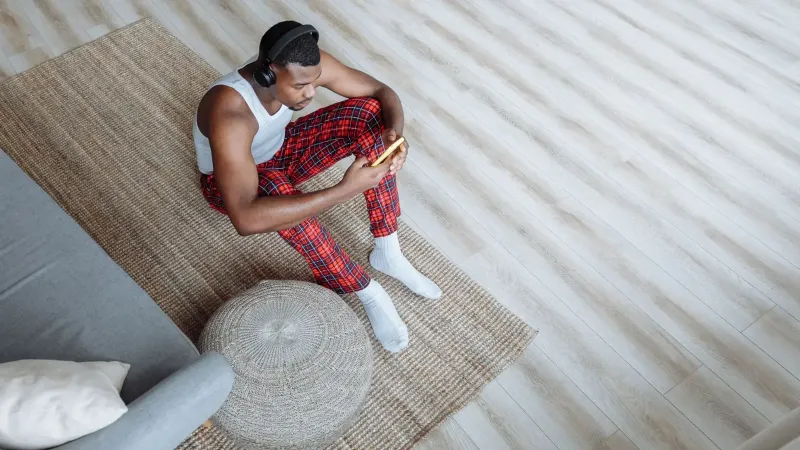 A man in plaid pajamas sits on the floor wearing headphones, using his smartphone in a cozy living room.