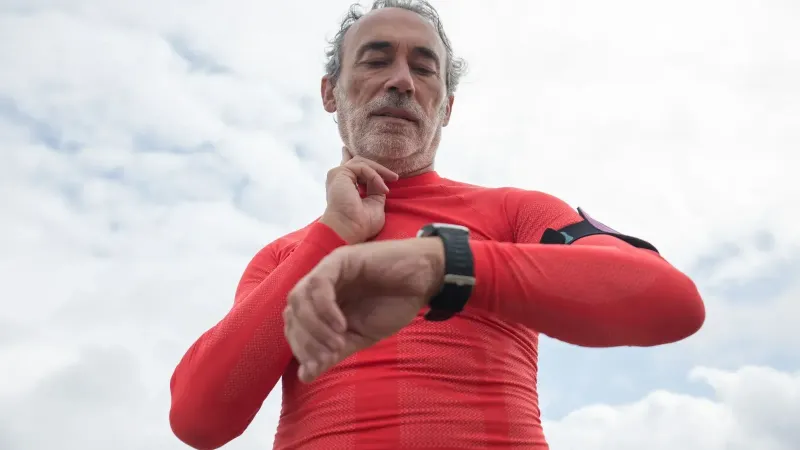Anciano en rojo desgaste activo comprobando reloj de pulsera al aire libre bajo el cielo nublado.
