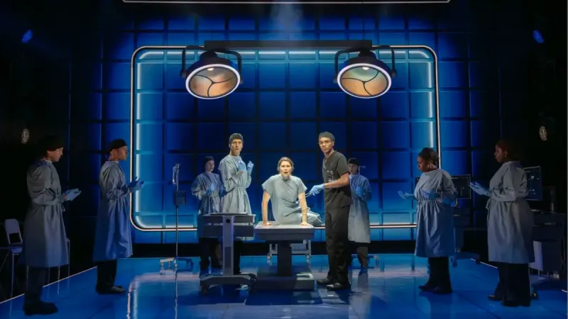 Three actors on stage in surgical gowns in an operation room.