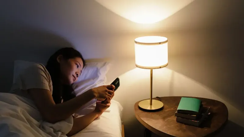 A woman relaxes in bed, using her smartphone beside a lit table lamp in a cozy room.