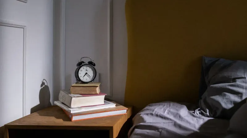 Un acogedor entorno de dormitorio con libros y un despertador en una mesita de noche junto a una cama.