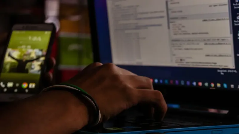 Close-up of a person working on a laptop and smartphone in a dimly lit office, showcasing remote work.