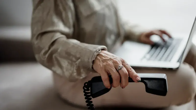 Una persona se sienta en un sofá escribiendo en una computadora portátil y sosteniendo un receptor de teléfono negro con la mano derecha.