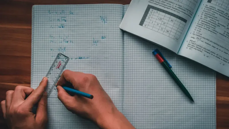 Una persona escribe ecuaciones matemáticas en un cuaderno usando una regla y un lápiz, enfocados en resolver problemas.