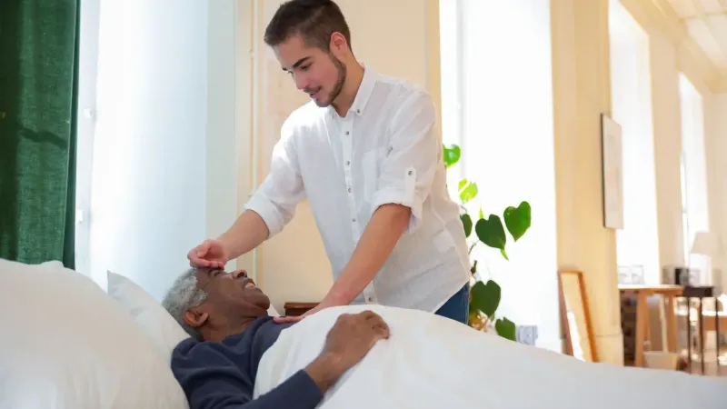 Un joven apoya a un anciano en la cama, ofreciendo cuidado en una habitación luminosa.