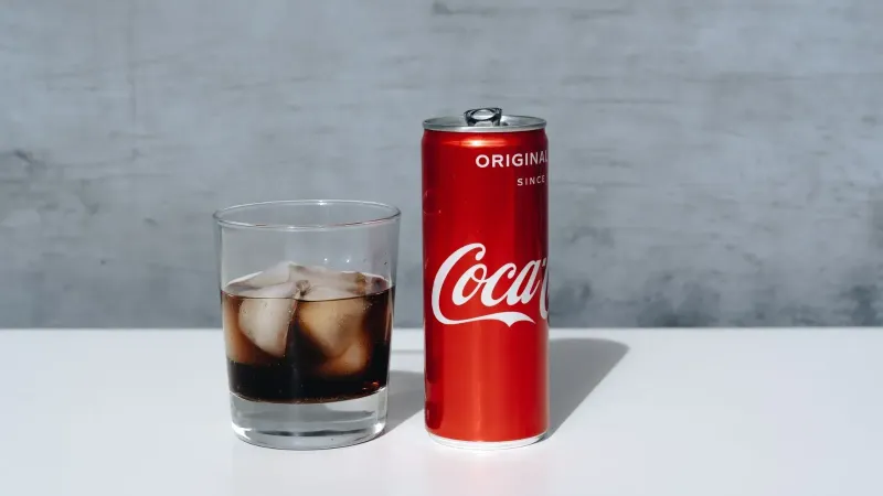 Lata de Coca-Cola refrigerada y beber con cubitos de hielo en vidrio sobre superficie minimalista.