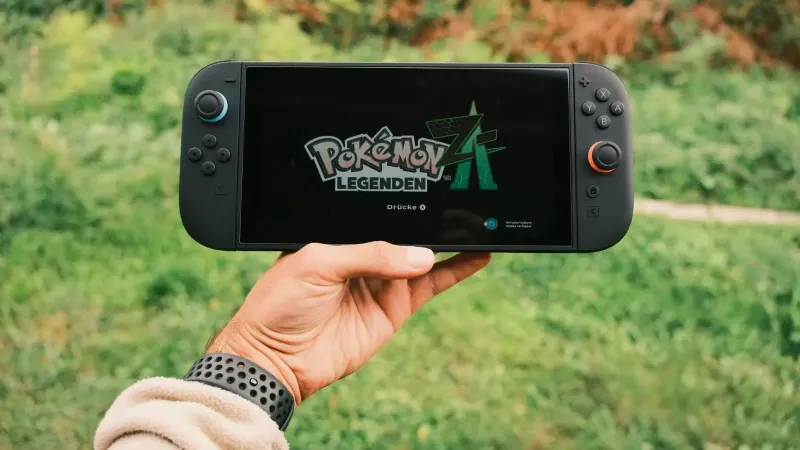 A hand holding a handheld gaming console displaying the Pokémon Legends game screen outdoors.