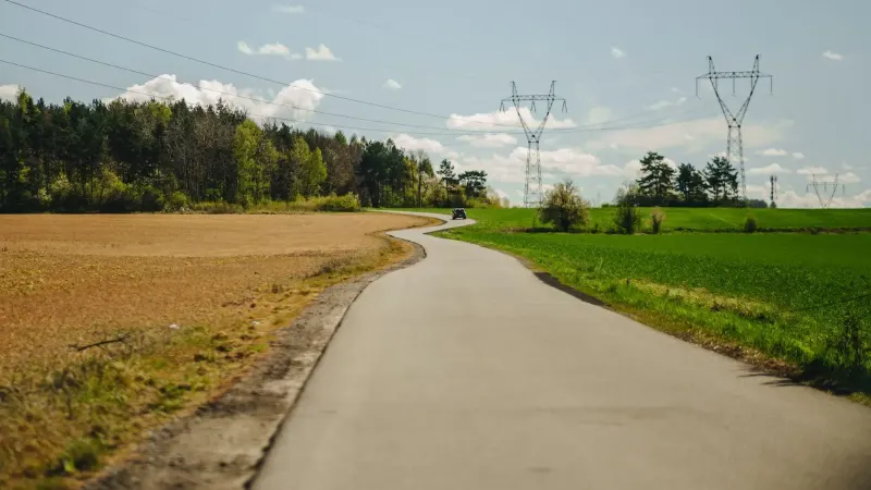 Camino curvo entre campos con líneas eléctricas y fondo forestal en un día soleado.
