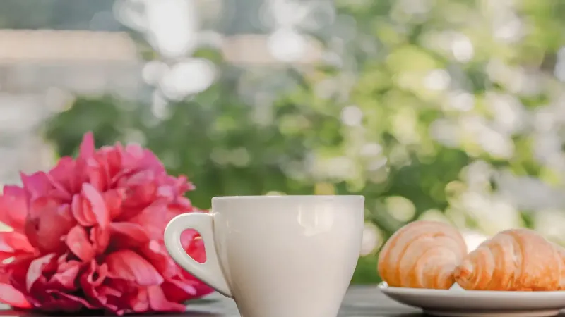 Taza de café caliente servido en mesa de madera con manojo de peonías rosas y plato de croissants recién horneados durante el desayuno en la terraza en la mañana soleada