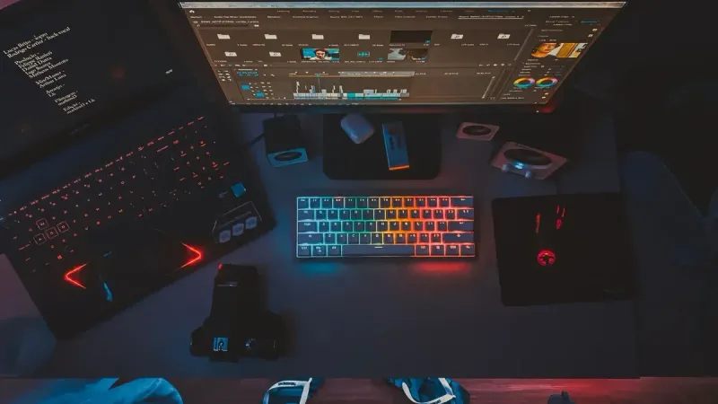 Un espacio de trabajo contemporáneo con monitores duales, un teclado iluminado con arcoíris y software de edición, perfecto para los entusiastas de la tecnología.