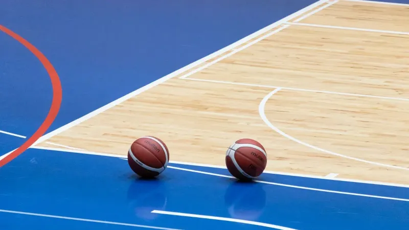 Two basketballs resting on a blue indoor basketball court with wooden flooring.