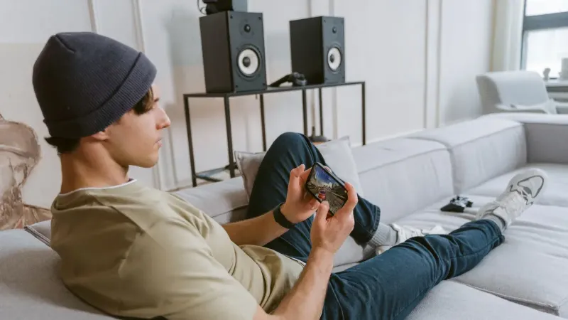 Joven casual jugando juegos móviles en el sofá con altavoces en el fondo.