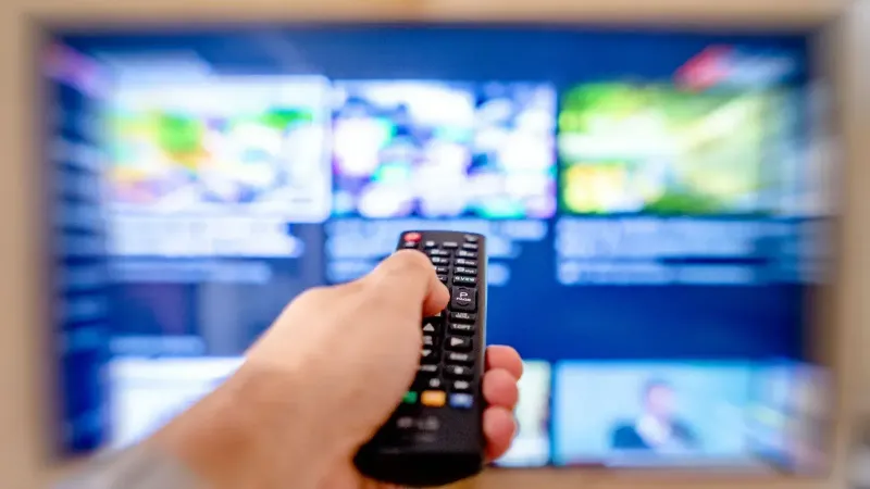 Hand holding remote control pointing at a blurred TV screen in a cozy indoor setting.