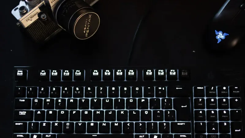 Una configuración tecnológica con poca luz que incluye un teclado iluminado, una cámara vintage y un mouse moderno.